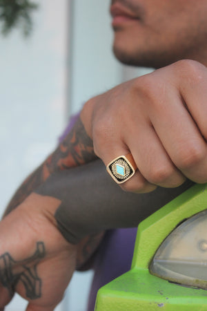 A close-up of a male model's pinky finger wearing a unique 14k Yellow Gold signet ring with a turquoise stone and engraved sunburst design.